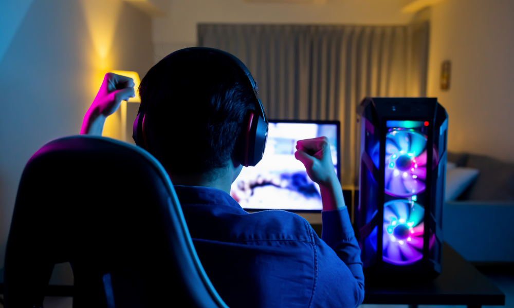 A male computer gamer wears a headset, raising his fists while looking at a PC monitor and setup that displays a video game
